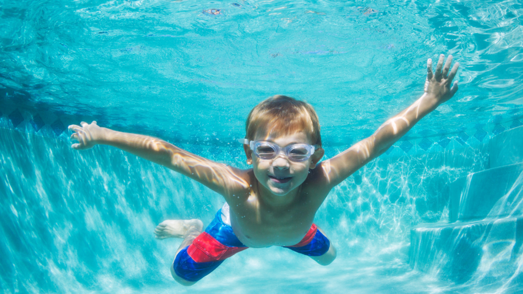 A young blonde boy with swim goggles and a blue and red swimsuit swims underwater