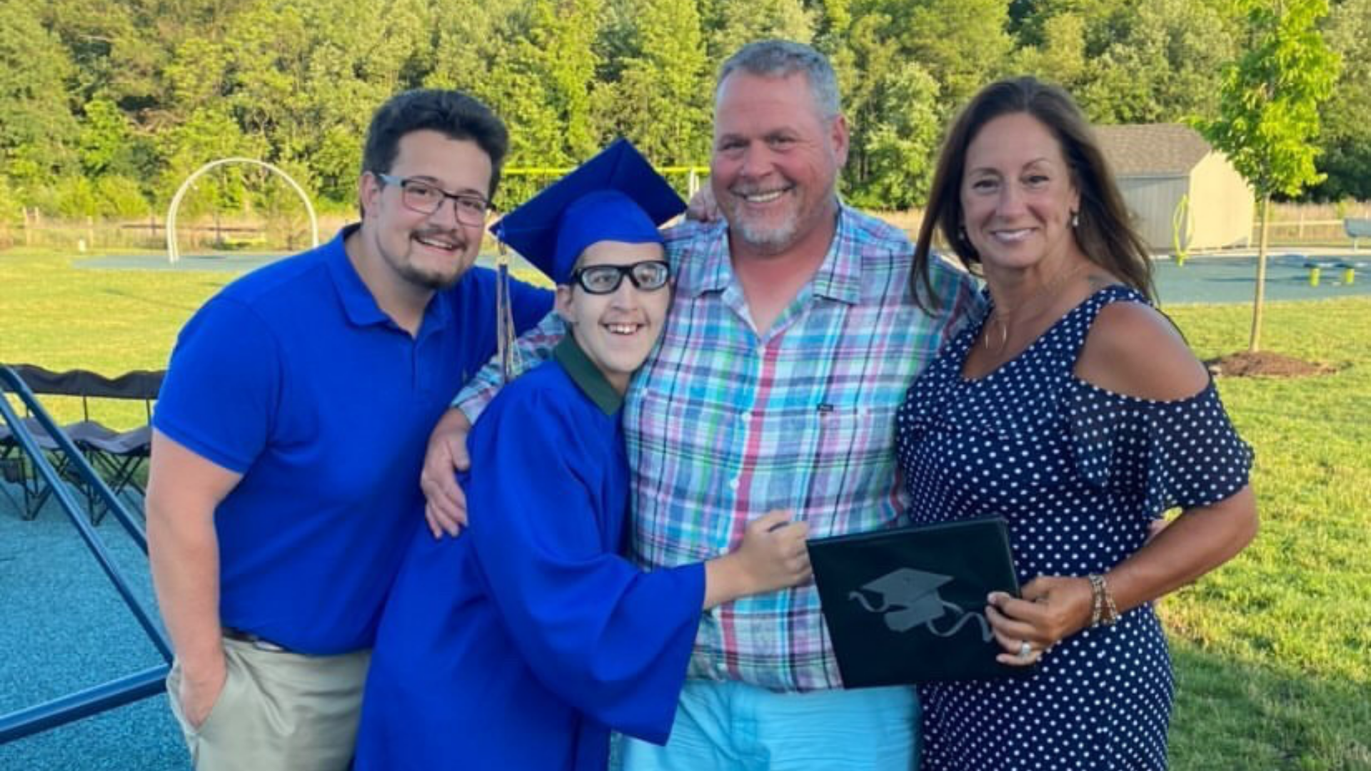 A graduate in a blue cap and gown and his three family members stand on an outdoor patio and smile