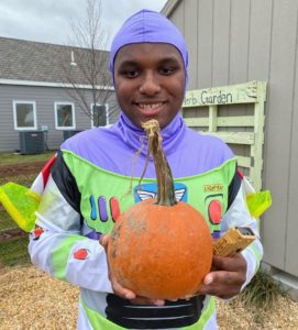 A young school aged boy is holding a pumpkin and smiling at the camera. He is wearing a Buzzlight Year costume for Halloween. 