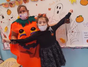 A preschool aged girl and her teacher are dressed up for Halloween. The teacher is wearing a giant pumpkin costume and the girl is dressed as a with in an all black dress. 