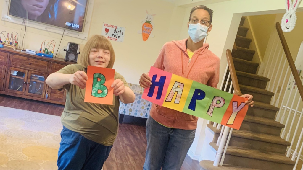 a female person served in a green shirt with blonde hair stands with a male paraprofessional in an orange sweatshirt and blue jeans holding signs that say "B Happy"