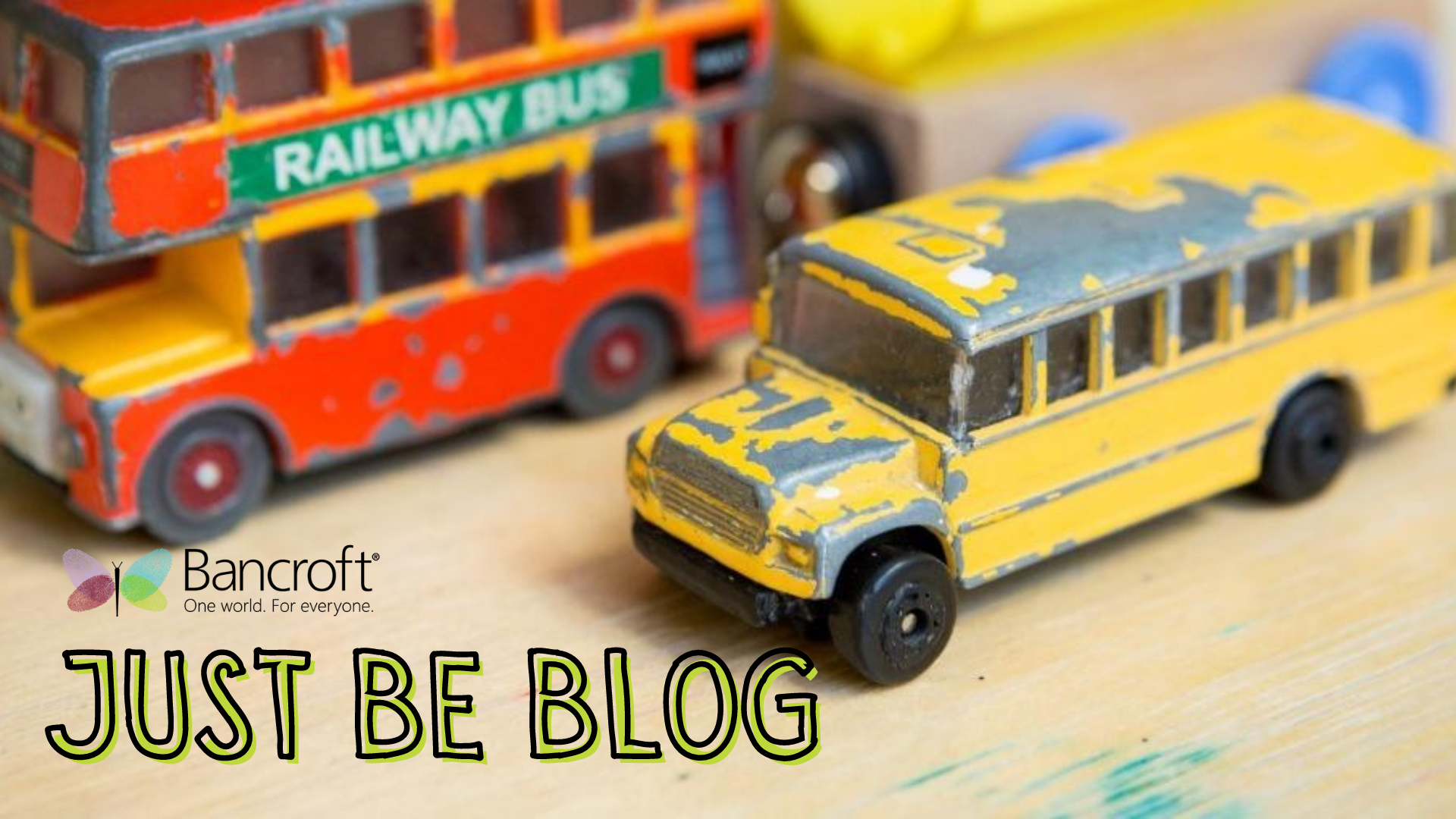 Toy busses, one a red double-decker bus and the other a yellow school bus, sit on a table with the caption "Just Be Blog" in yellow letters with a black outline
