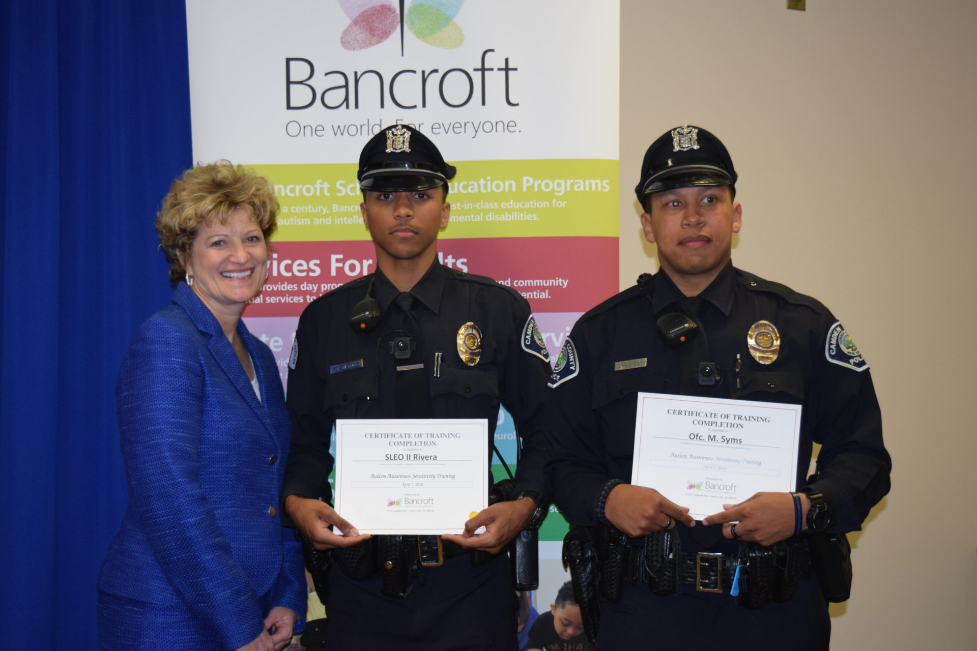 A light skinned woman stands with two dark skinned police officers. They offers are on her right and they are holding certificates.