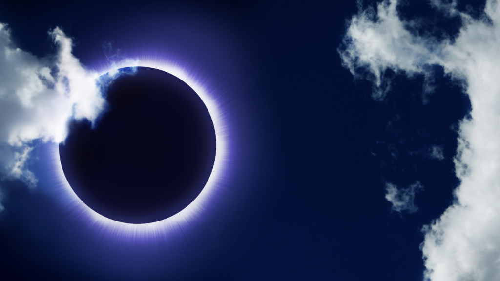 an eclipse; the sun covered by the dark moon in a dark blue sky with white clouds