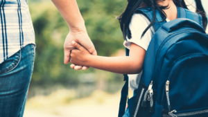 Student with black hair, a white shirt, and a blue backpack holds their parent's hand as they walk to school