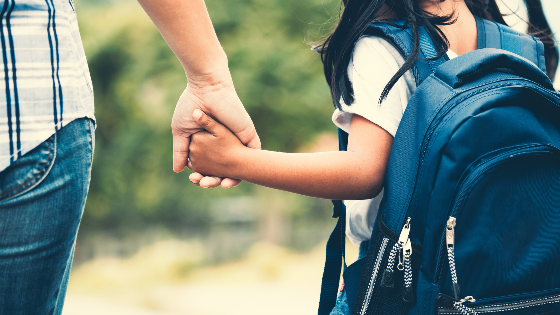 Student with black hair, a white shirt, and a blue backpack holds their parent's hand as they walk to school