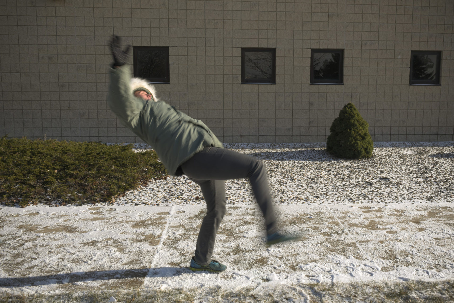 A person in a green jacket and gray pants slips on an icy sidewalk