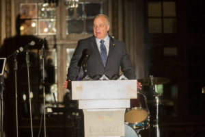 light-skinned man in a gray suit with a blue shirt and tie stands at a Bancroft podium talking into multiple microphones