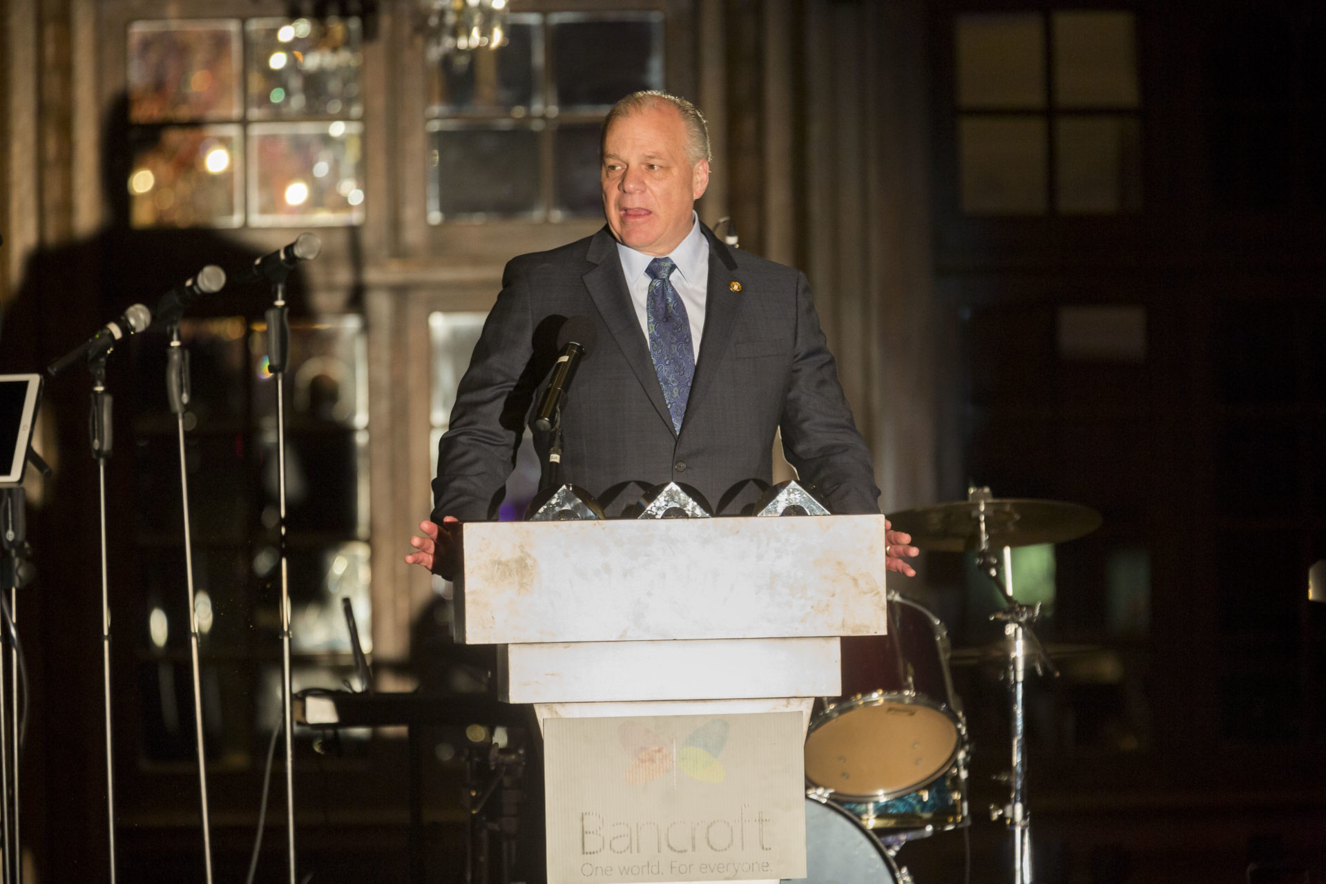 light-skinned man in a gray suit with a blue shirt and tie stands at a Bancroft podium talking into multiple microphones