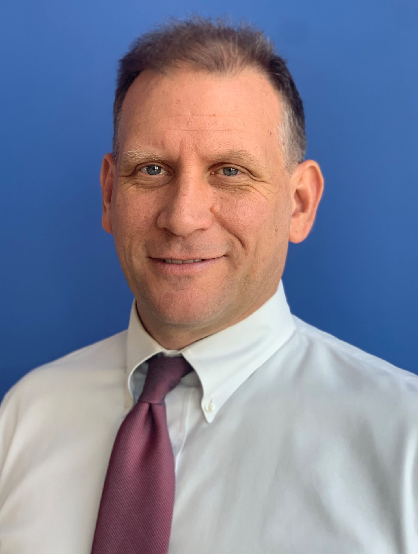 Pat Senft, a light-skinned man with short gray hair in a white button down shirt and purple tie, stands in front of a blue background and smiles