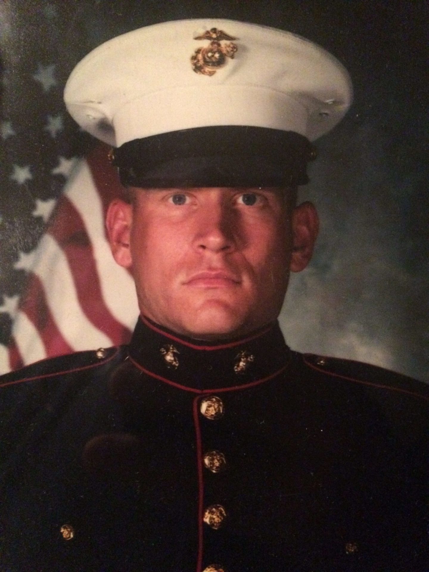 Pat Senft, a light-skinned man wearing a military outfit, poses in front of an American flag background