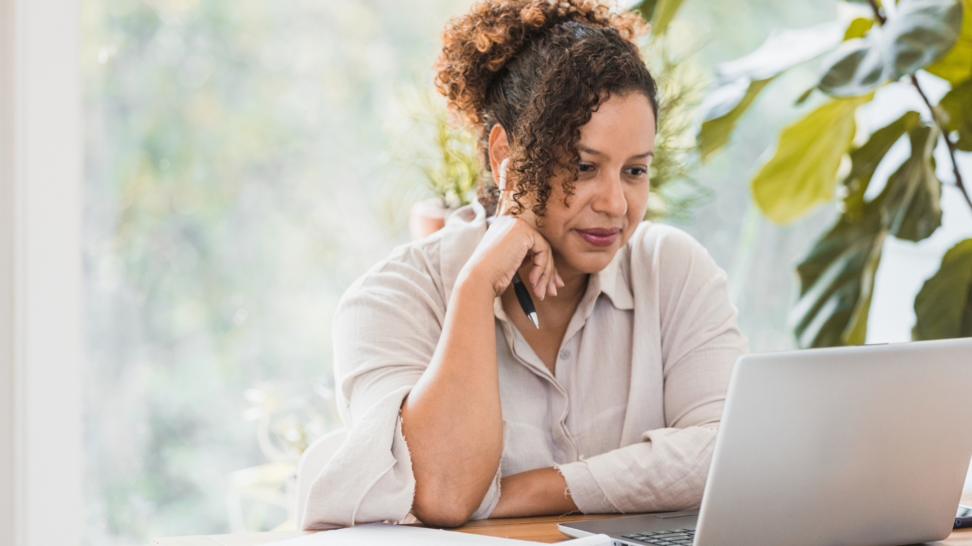 woman with brown curly hair in a cream button down shirt sits in front of plants at a wooden table staring at a silver laptop with her head on her hand holding a pencil