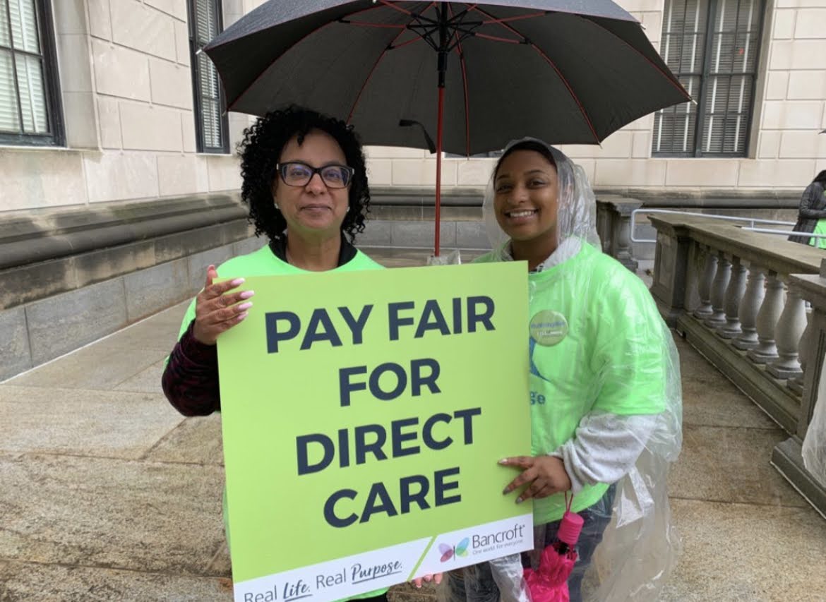 two woman stand outside in neon green shirts holding a black umbrella and a sign that says "Pay Fair For Direct Care" and features Bancroft's real life real purpose logo