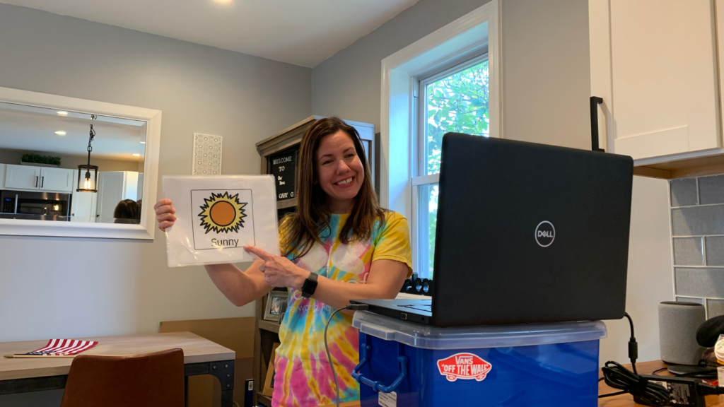 woman in a tiedye shirt holds a photo with a sun on it that says "Sun" in front of a gray Dell laptop on top of a blue bin in a house