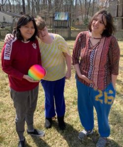 three young women stand in a grassy backyard smiling as one of them holds a medium-sized multi-colored ball
