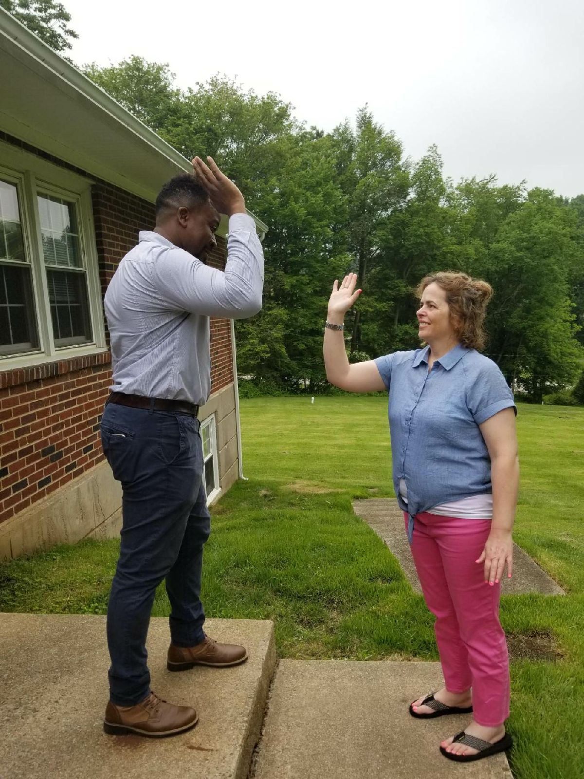 a man in a blue shirt and pants stands on a patio and high fives a woman in a blue button down shirt and pink pants