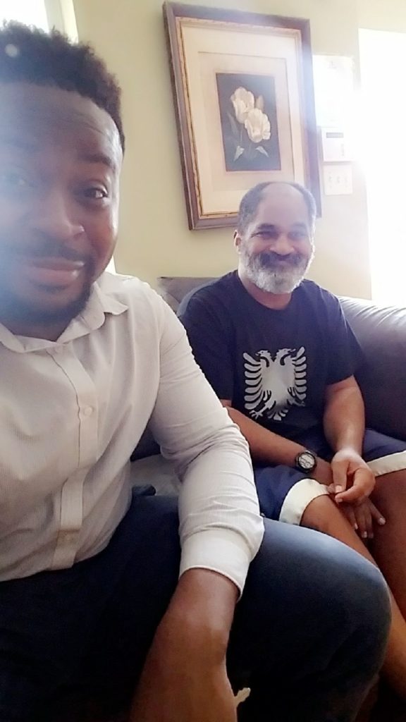 a man wearing a white button down shirt and a man wearing a black t-shirt sit on a couch and take a selfie
