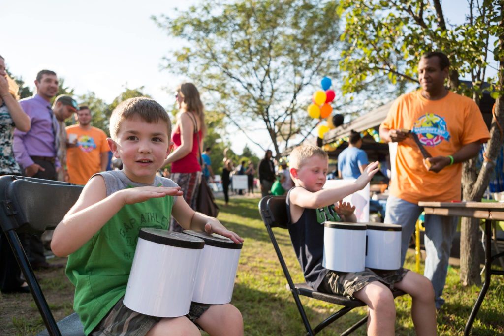 two little boys are sitting on chairs playing bongos while dozens of people stand around them socializing