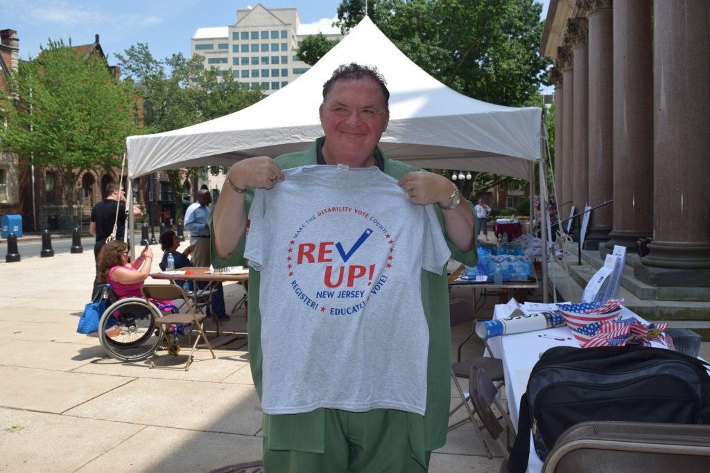a man in a green shirt stands holding a gray Rev Up New Jersey voting t-shirt