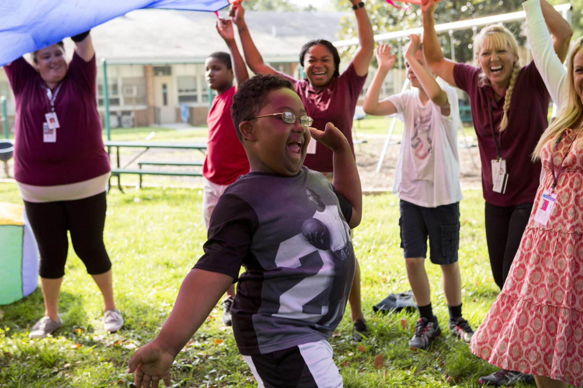 A young boy is running under a parachute. He has his arms spread out wide on his sides and is surrounded by Bancroft staff.