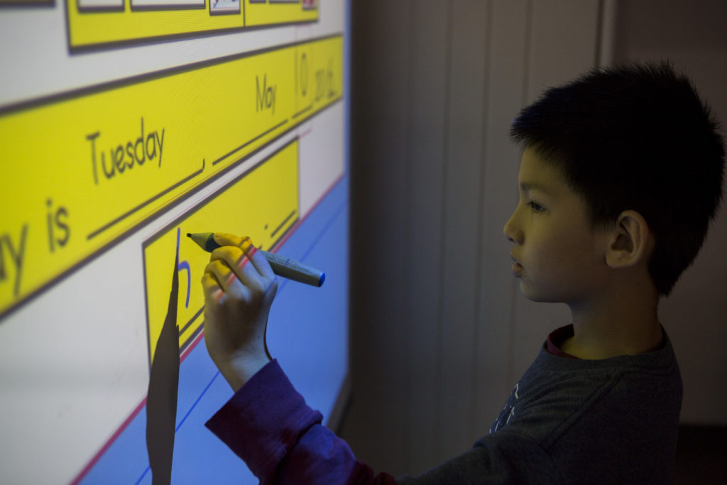 a young boy in a blue shirt stands at at a smart board and writes