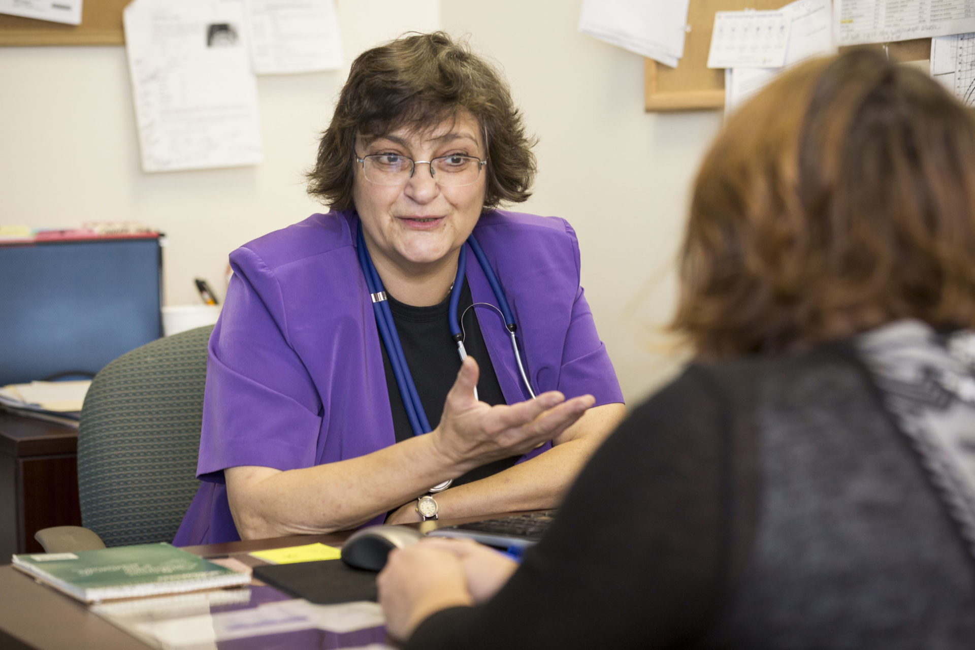 a woman with short black hair wearing a purple lab coat and stethoscope sits behind a desk talking to a woman with short brown hair in a black shirt