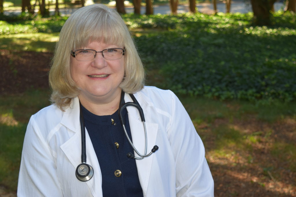 Lisa Alberts; a light-skinned woman with blonde hair and blue eyes wearing a white lab coat, blue top, and stethescope