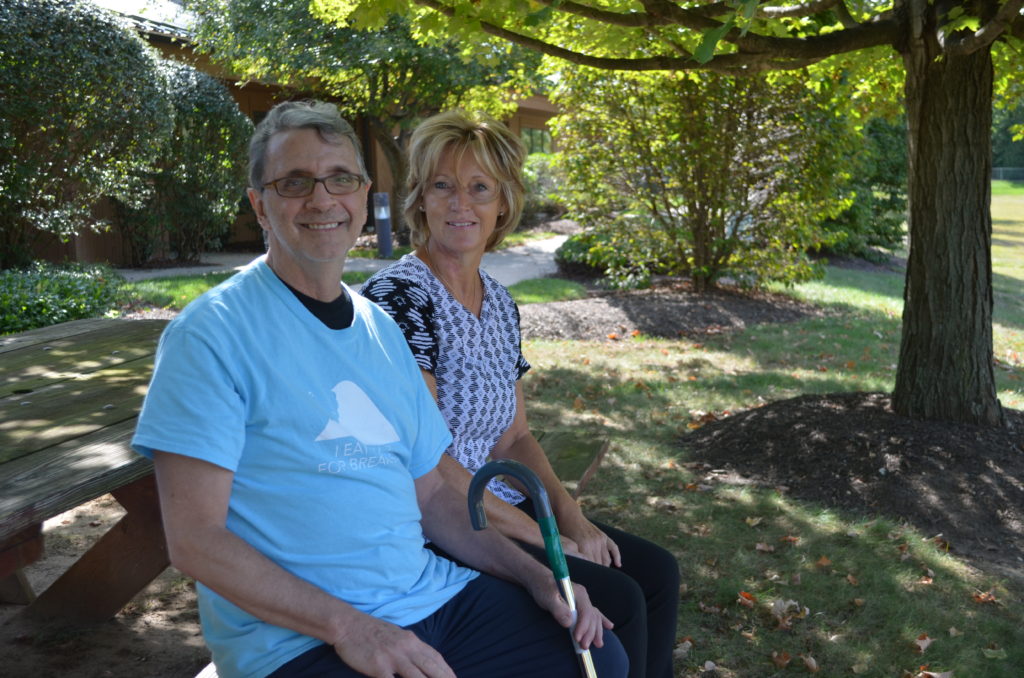 a man in a blue shirt and glasses sits at a wooden table with a woman in a blue and white patterned shirt in a woody area