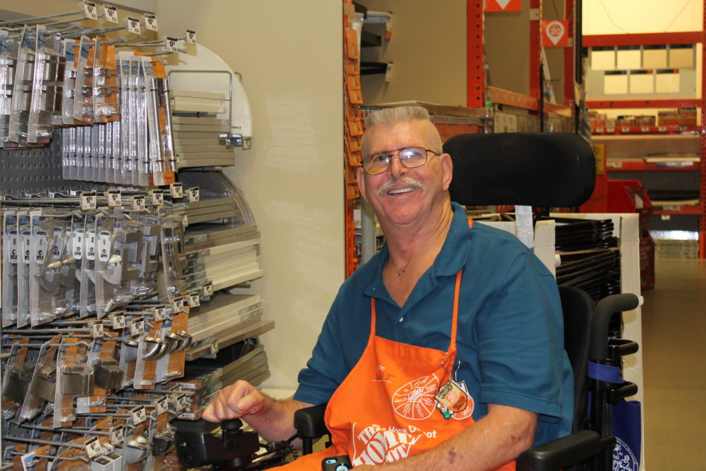 a man sits by a shelf in a hardware store wearing a blue shirt and "Home Depot" apron