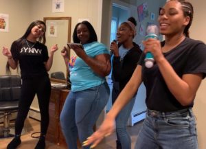 four young women stand inside a living room singing into microphones and smiling