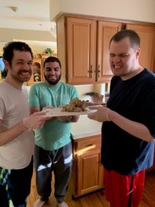 three young men stand inside a kitchen holding a plate of homemade cookies and smiling