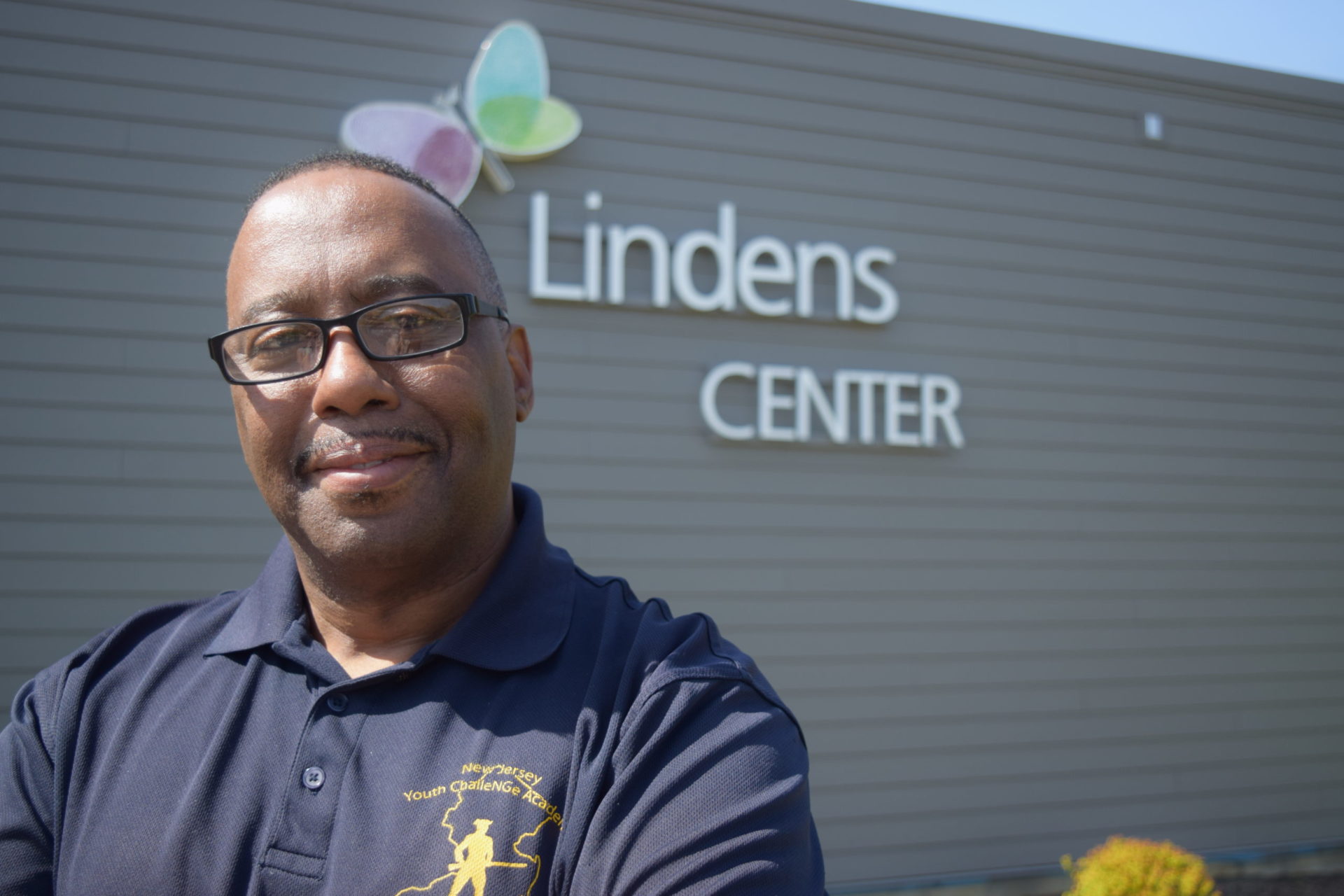 Michael Bradley, a dark-skinned man in a blue polo, stands outside of the Lindens Center building and smiles