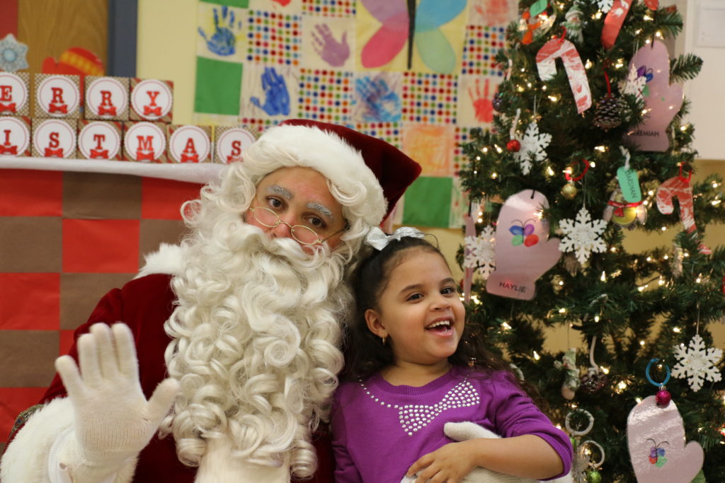 a little girl in a purple shirt with a gem bow sits on Santa's lap in front of a decorated Christmas tree