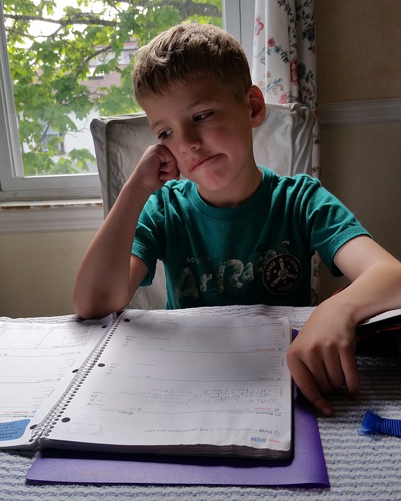 a little boy in a teal shirt sitting in front of an open notebook holds his head against his hand looking bored