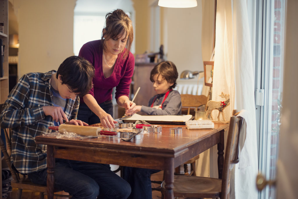 a mother and two boys sit and stand at a table making cookies