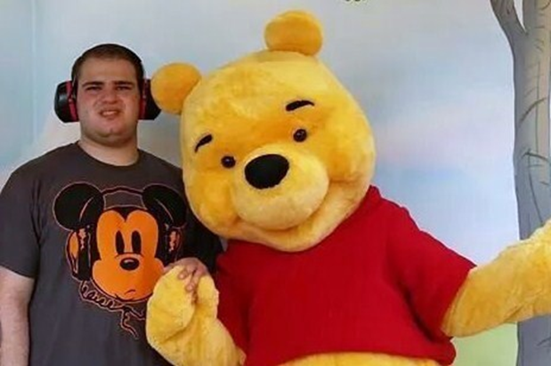 a person served wearing headphones and a gray mickey mouse t-shirt stands with a life-sized Winnie the Pooh, a yellow bear in a red t-shirt