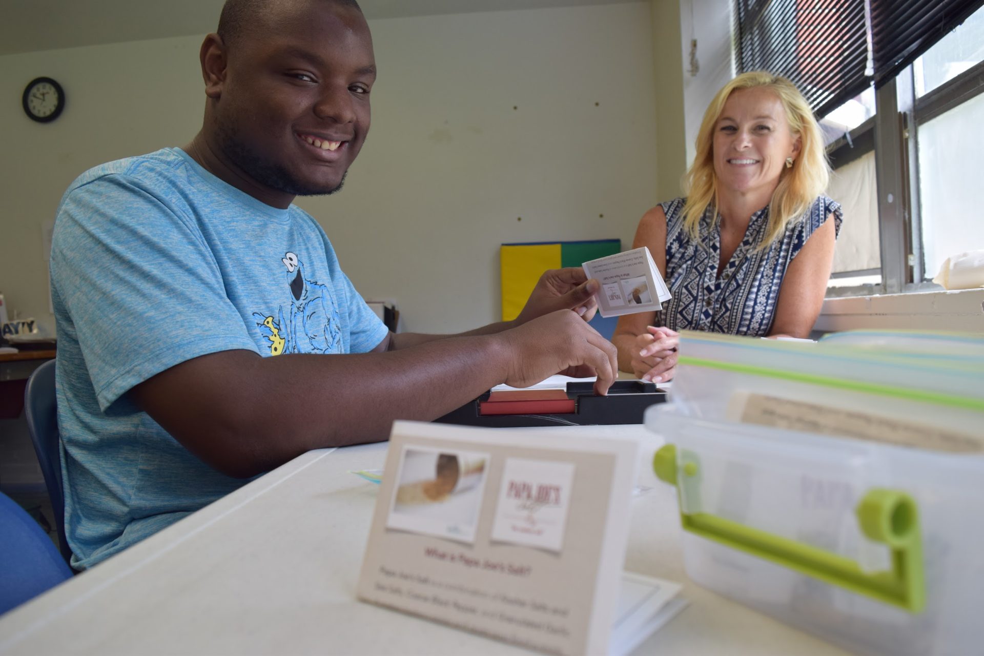 a person served sits in a blue cookie monster shirt holding a card out to a female paraprofessional in a blue top, smiling