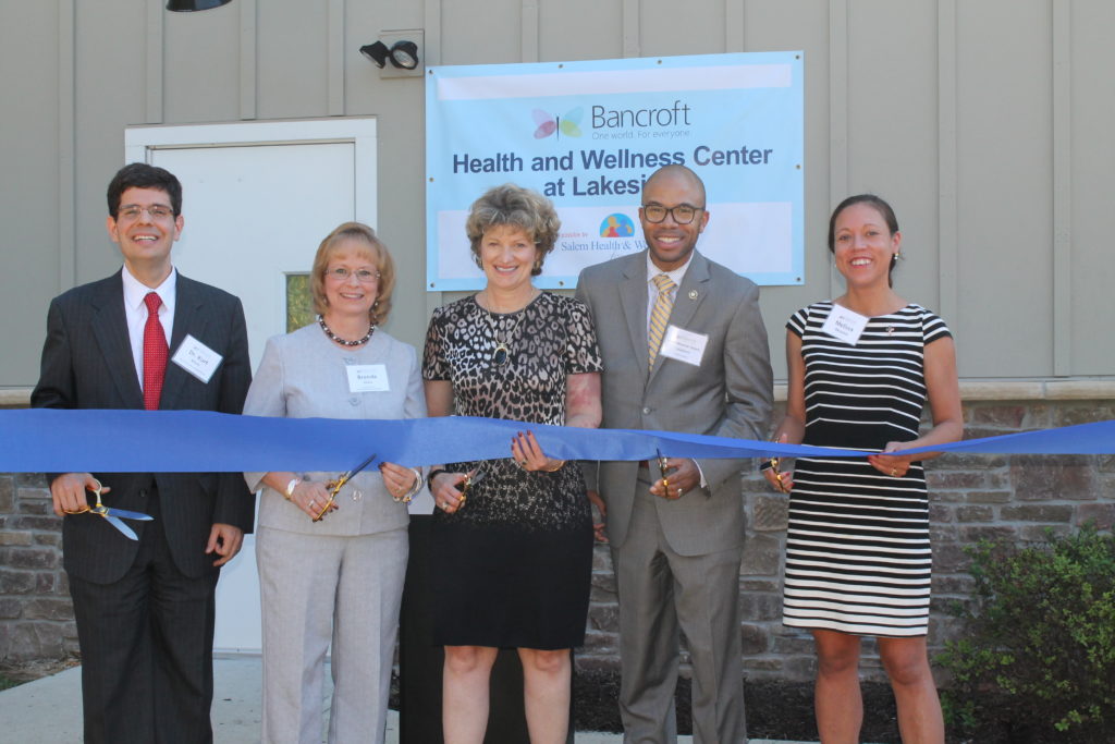 five Bancroft board members stand holding a blue ribbon and large scissors outside of the new Health and Wellness Center at Lakeside building