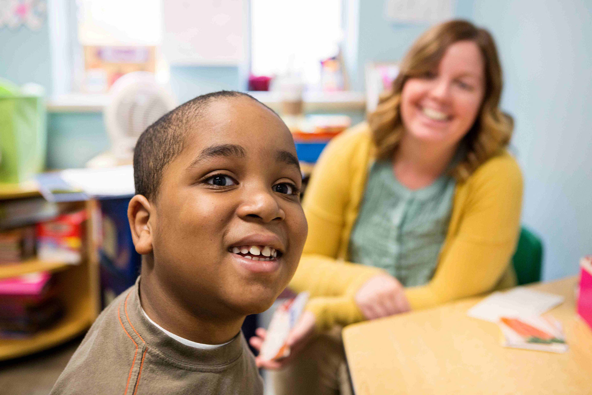 a young boy in a brown shirt sits at a table and smiles while a female paraprofessional in a yellow cardigan watches him, smiling