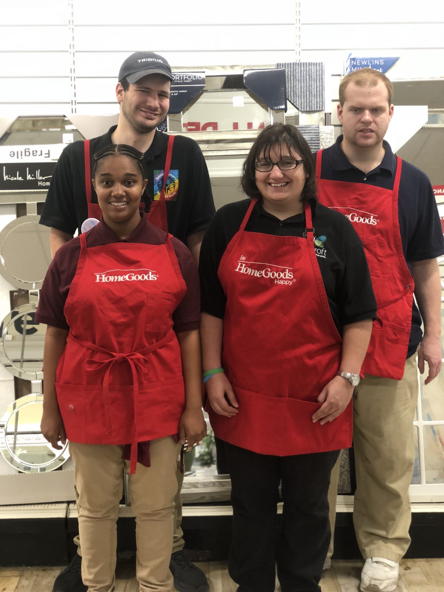 four people in a red Home Goods aprons stand against the shelf in a Home Goods store and smile