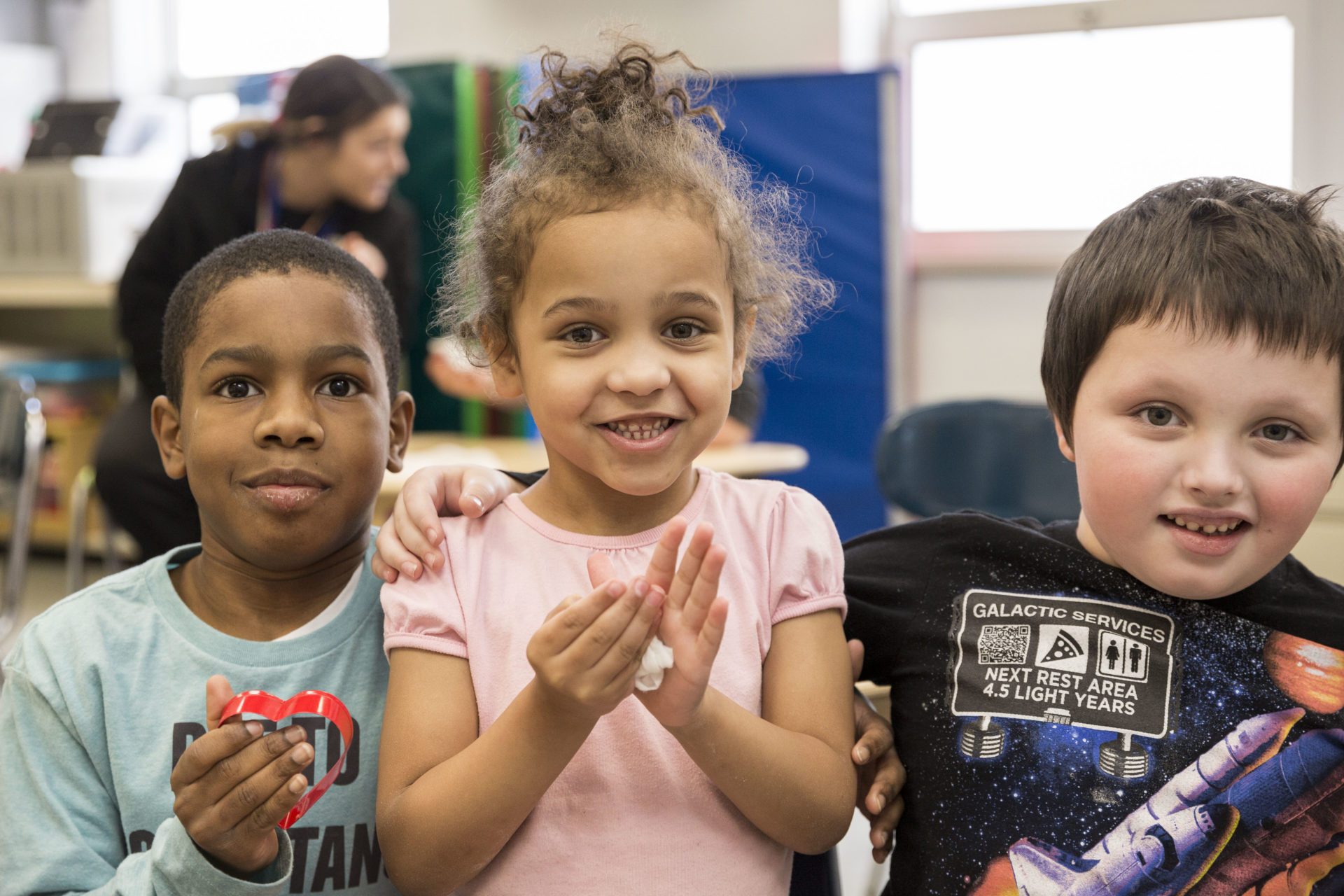 Three preschool students, two boys and one girl posing together for a photo. The girl is wearing a pink shirt and has brown hair, the boy on the right is wearing a black spaceship shirt and the the boy on the left is wearing a light blue shirt.