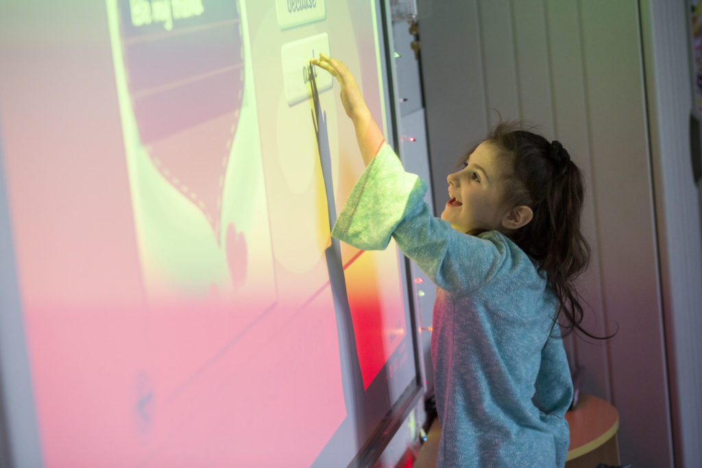 Preschool aged girl with brown hair pointing at a smart board screen which is lit up pink.