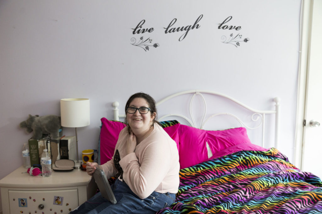 A girl sits on her bed holding her iPad. Her bedding is colorful and her room is bright.