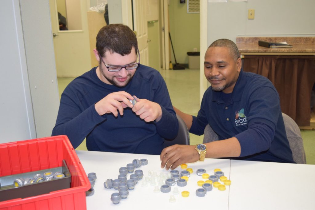 A male student in a blue shirt and male paraprofessional in a blue shirt smiling playing with caps and bottles
