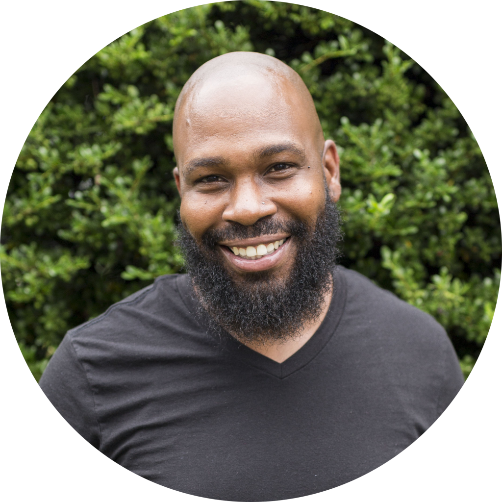 Male direct support professional smiling and posing for the camera. He is wearing a black t-shirt and is standing in front of a tree.