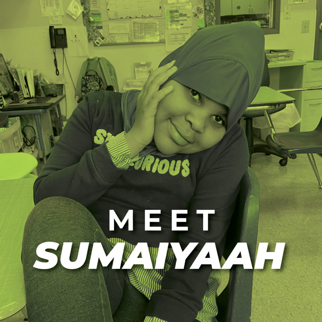Sumaiyaah, a young girl with a purple shirt and leggins sits at a table in a classroom holding her hand against her face
