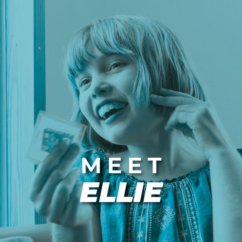 Ellie; a young girl with short hair and patterned shirt sits at a table holding a card and smiles