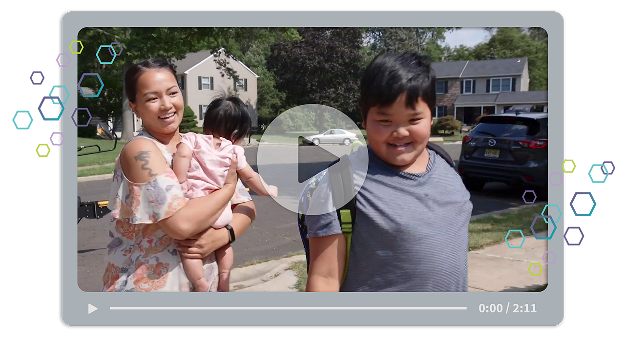 a young boy in a gray shirt wearing a backpack stands with a woman in a floral top holding a baby in a neighborhood