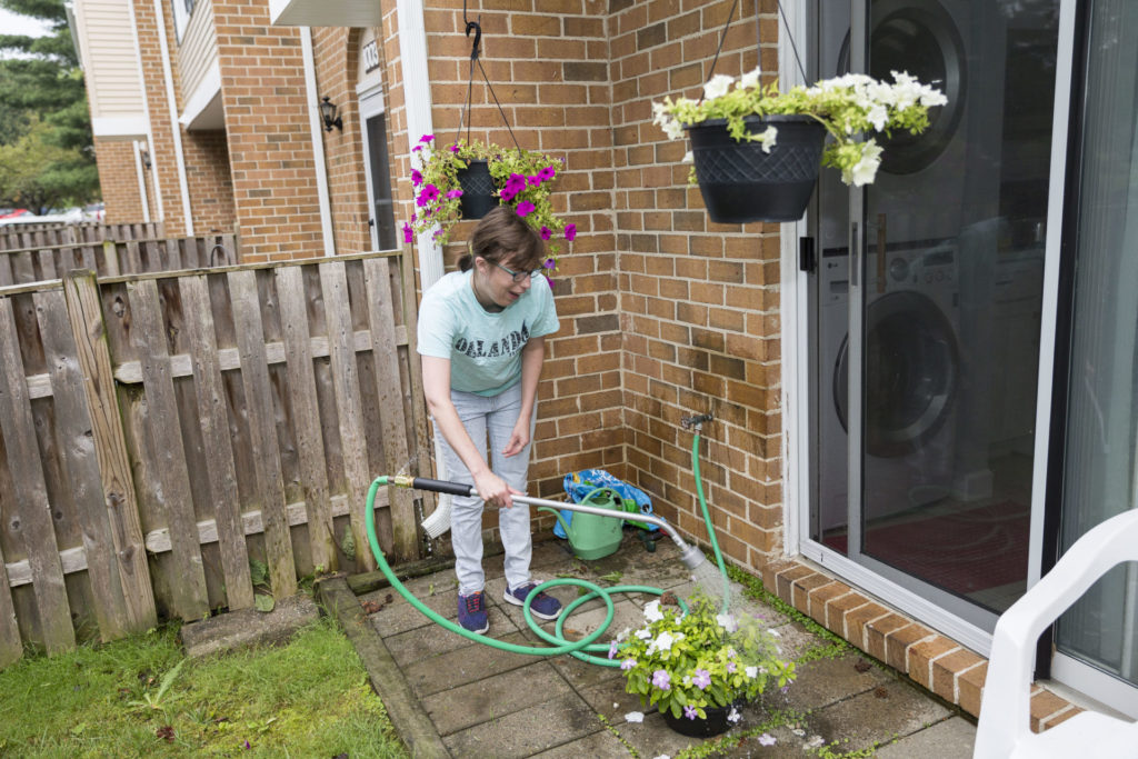 An adult woman outside of her home watering plants and flowers with a green hose.