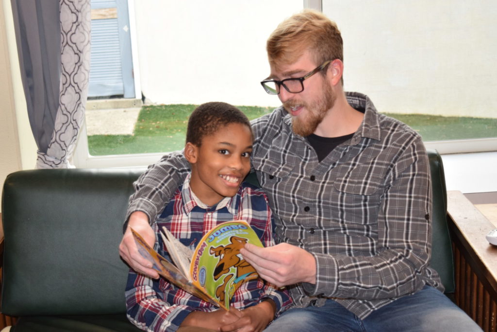 A young boy sitting on a couch reading a book, he is wearing a red plaid shirt. There is a paraprofessional sitting next to him, holding the book. He is also wearing a plaid shirt but it is grey.